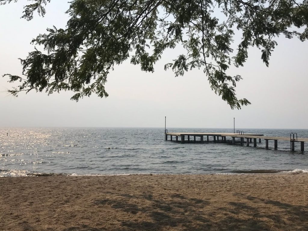 A photograph of a West Kelowna beach during the daytime taken September 8 2020. The sky is visibly smokey, completely grey with no visible horizon. The sunlight reaching the beach looks darker than normal and red-shifted because of the smoke.