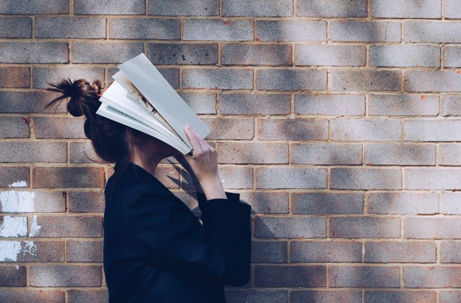 Woman with Book in front of natural rustic red brick background holding book up to her face.