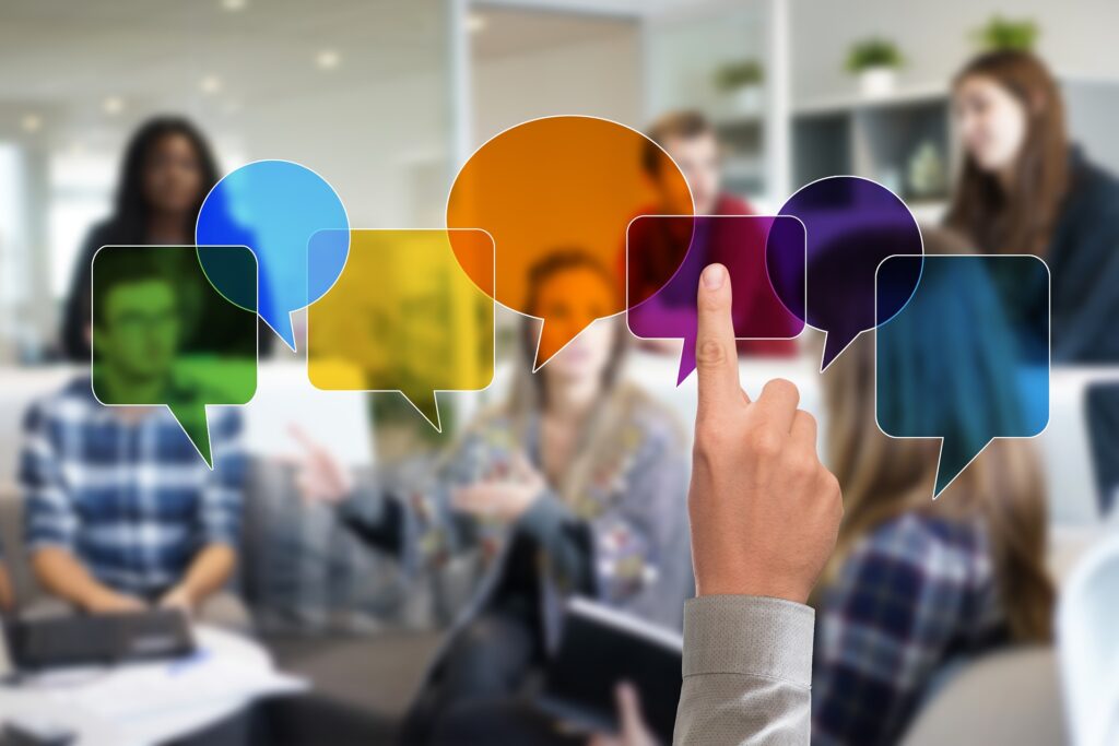 A line of overlapping, colourful speech bubbles is overlaid over a slightly blurred image of a group of people talking. A hand reaches up from the bottom of the image, pointing at one of the speech bubbles.