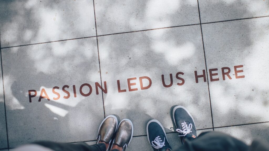 Top down view of a concrete path with the text "Passion Led Us" written on it. Two people's feet can bee seen surrounding the text.